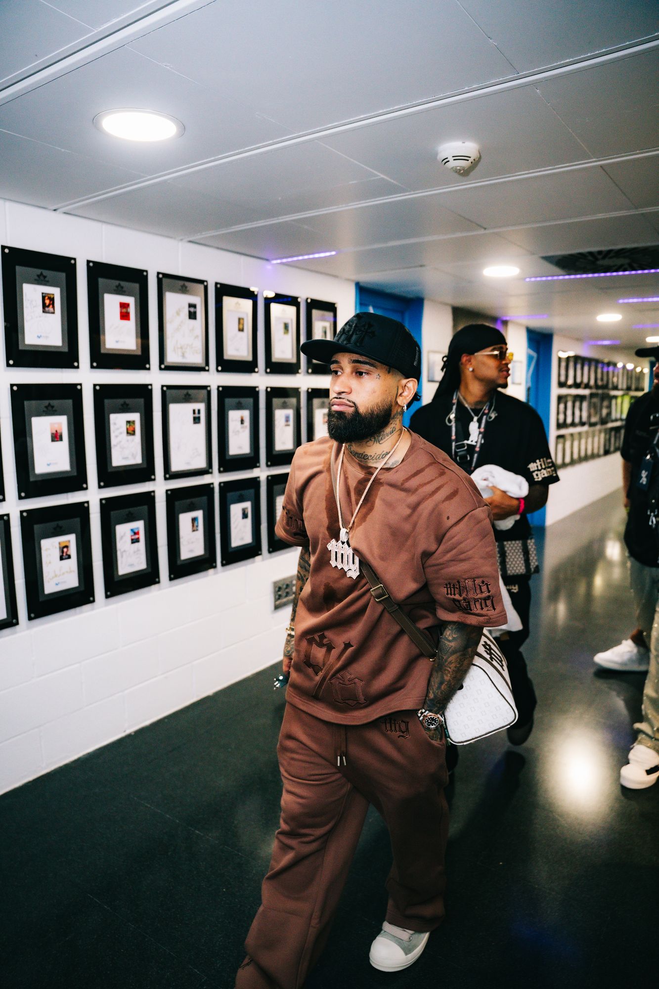 Bryant Myers in a corridor at Movistar Arena wearing the brown Millo Gang set and carrying the monogram bag